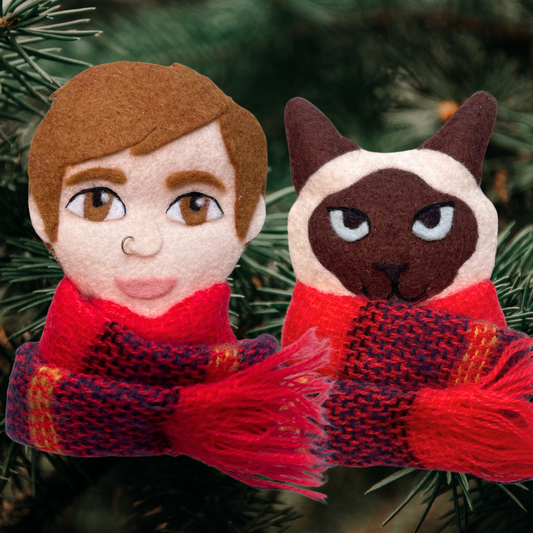 Felt figurines of a person and a cat wearing scarves against a green foliage background